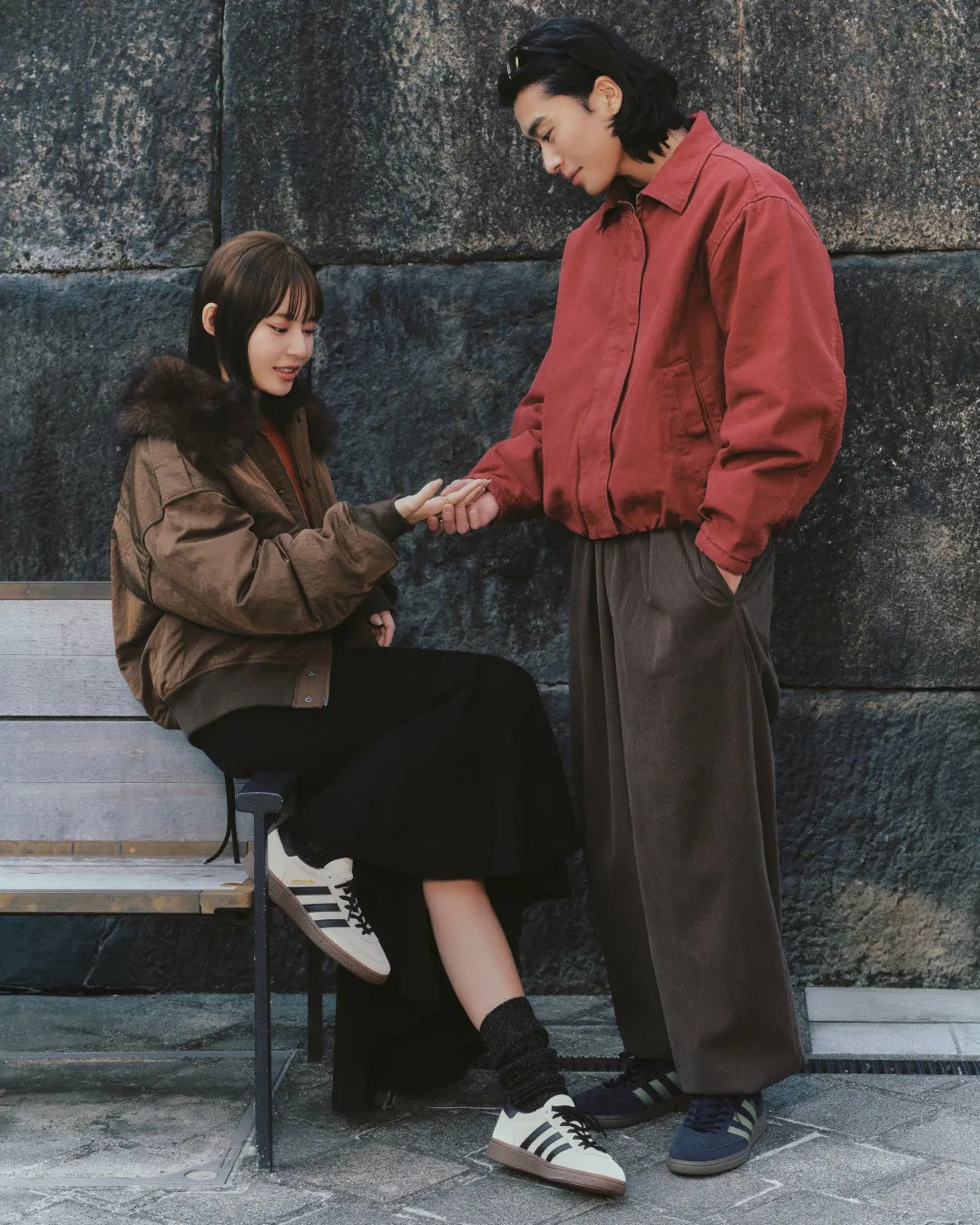 公園のベンチに座る女性と、彼女に手を差し伸べる男性が写った写真。二人はカジュアルな服装で、穏やかな表情を浮かべている。背景には石壁があり、落ち着いた雰囲気の情景である。