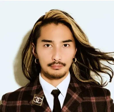 長髪の男性が、チェック柄のスーツとネクタイを着用し、イヤリングとブローチをつけている写真。