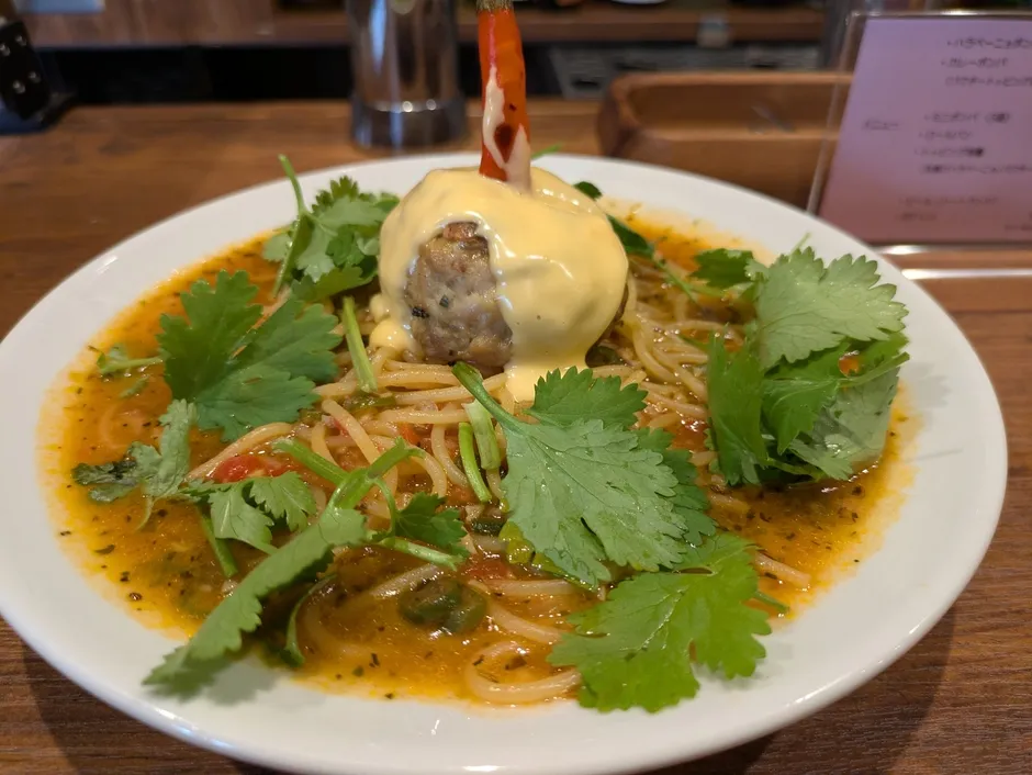 白い皿に盛られたパスタ料理。大きなミートボールが中央にあり、チーズソースがかかっている。香菜が添えられており、パスタはトマトベースのソースに浸されている。メニュー表も見える。