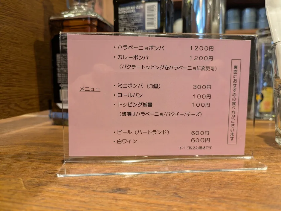 お店のメニュー表の写真です。ハラペーニョボンバ、カレーボンバ、ミニボンバ、ロールパン、トッピング増量、ビール、白ワインなどのメニューと価格が記載されています。