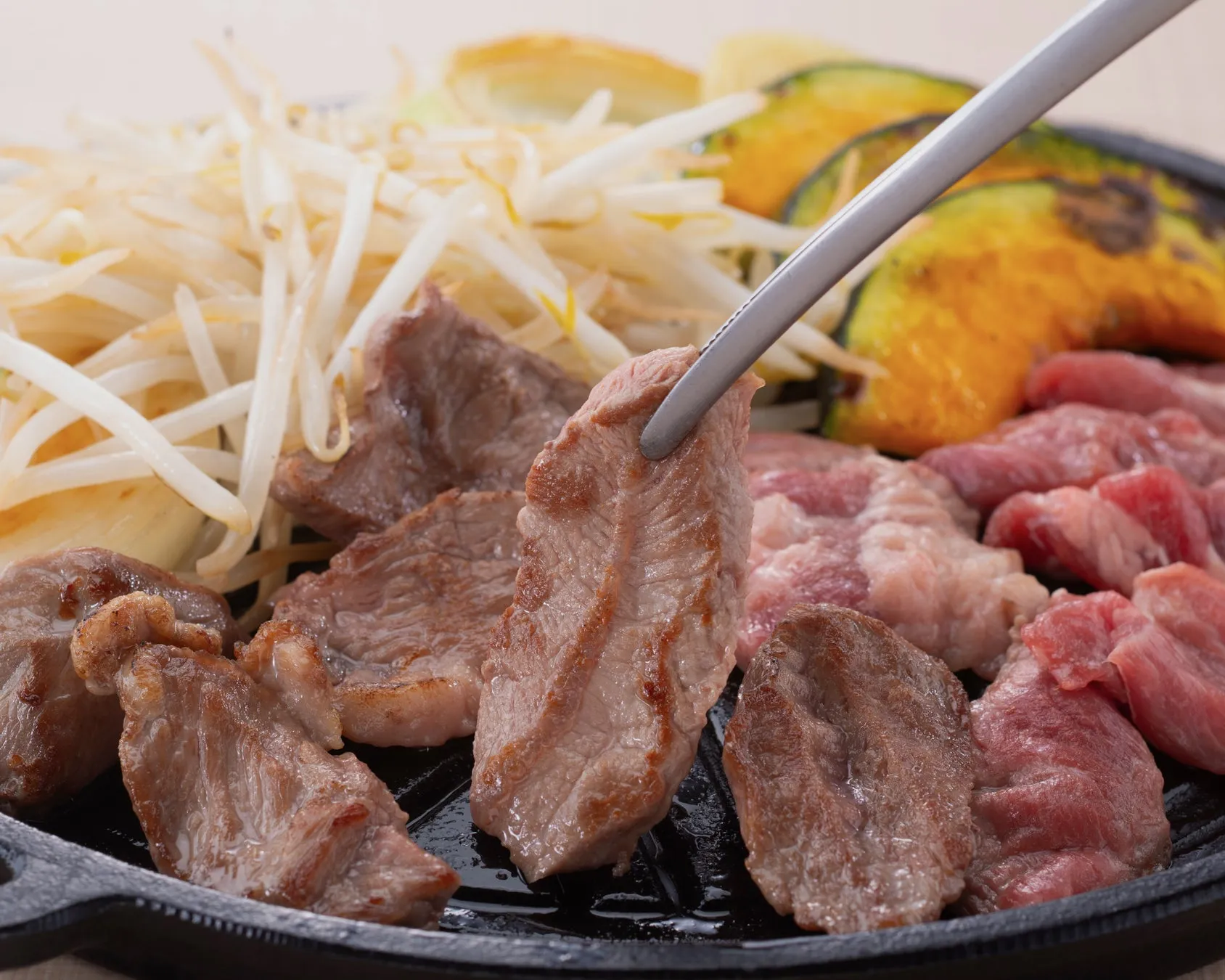 焼肉のクローズアップ写真。鉄板の上で焼かれた肉、もやし、カボチャが写っています。トングで肉をつまんでいる様子も捉えられています。