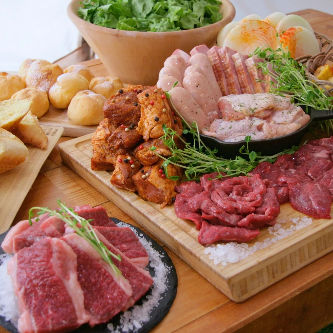 木製のテーブルの上に、様々な種類の肉、野菜、パンが並べられた美味しそうな料理の写真です。焼く前の肉、ソーセージ、鶏肉、野菜、パンが木製のまな板や皿に盛り付けられており、食欲をそそります。