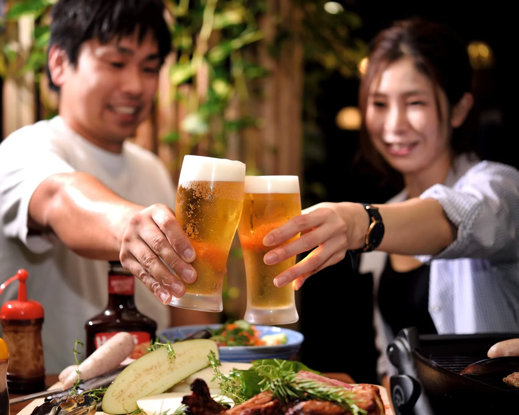 二人の人がビールグラスを乾杯している写真です。テーブルにはグリル料理や野菜が並んでいます。
