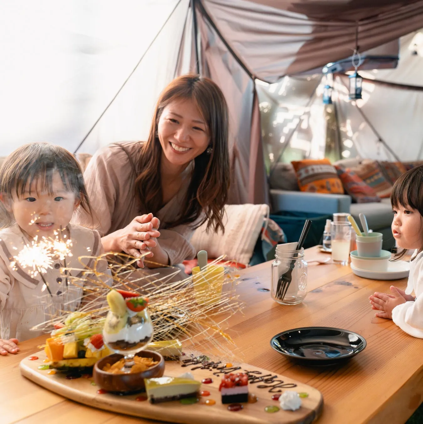 テーブルを囲んで楽しそうにしている家族の写真。子供たちは花火に興味を示し、母親は笑顔でそれを見守っている。テーブルには美味しそうな料理が並べられ、特別な日のように見える。