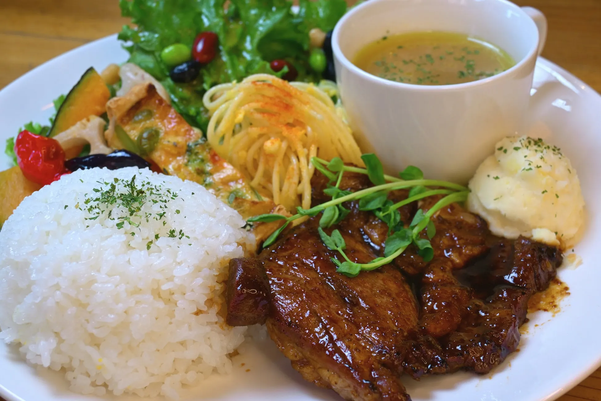 白い皿に盛られた、ステーキ、ご飯、スープ、パスタ、サラダなどの料理のクローズアップ。