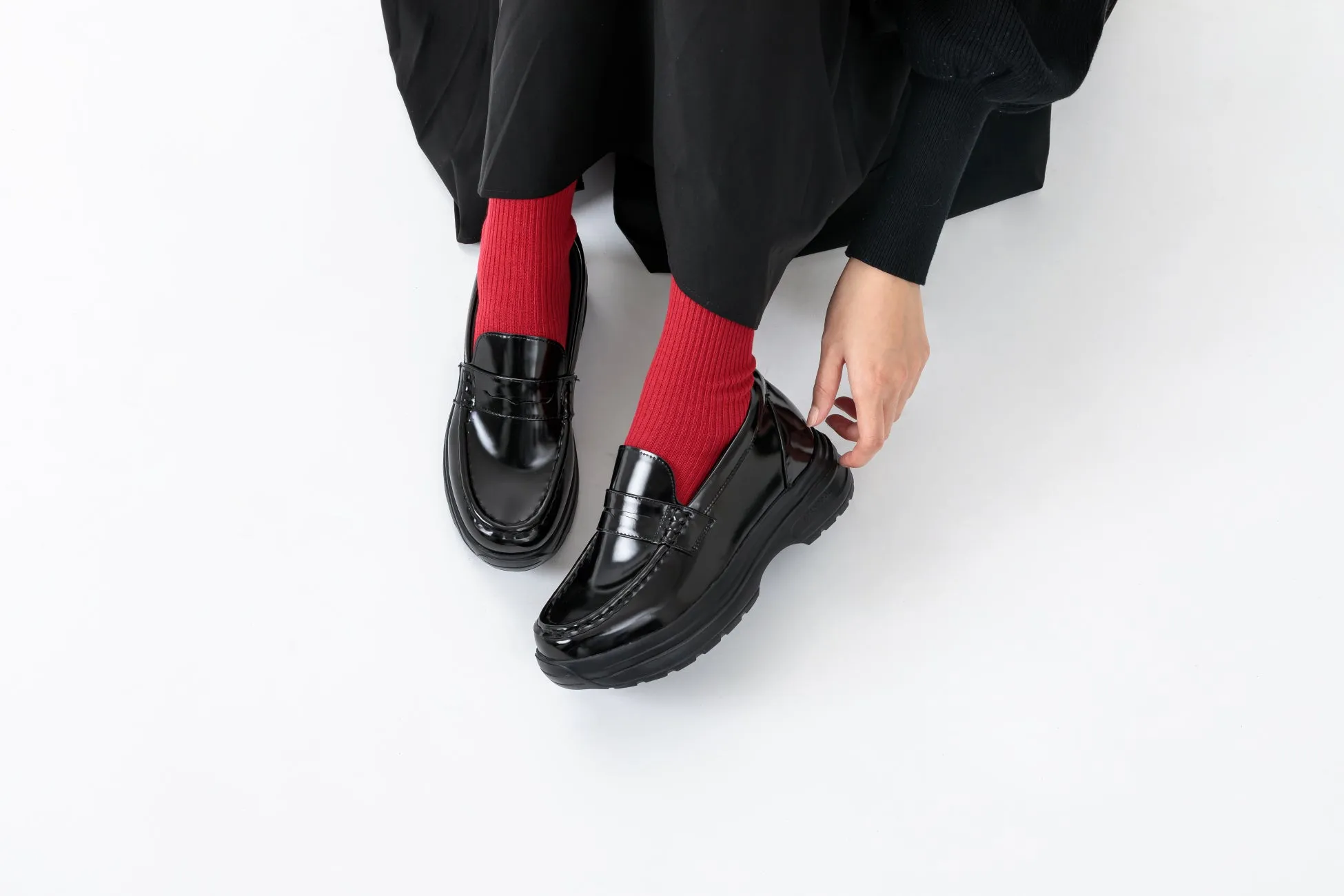 黒いローファーと赤い靴下を履いた人の足元の写真。人物はローファーに触れている。