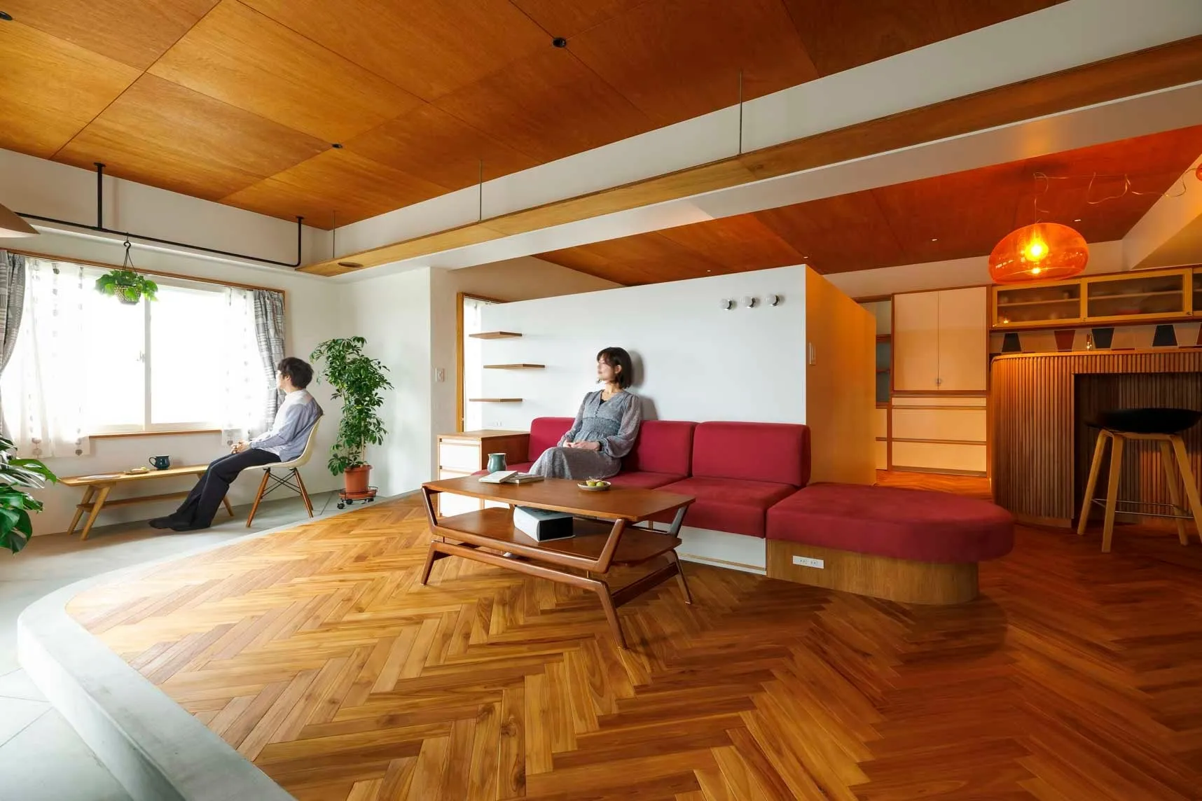 木材を基調としたモダンなリビングルーム。窓から光が差し込み、二人の人物がくつろいでいる様子が描かれています。赤いソファと、ヘリンボーンの床が印象的です。
