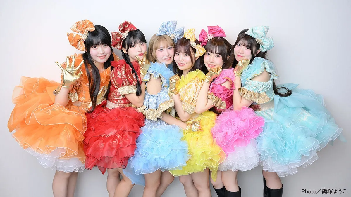 カラフルな衣装を着た7人組の女性アイドルグループの写真です。各メンバーは、オレンジ、赤、青、黄、ピンク、水色などの異なる色のドレスを着ています。全員がカメラに向かって微笑んでいます。