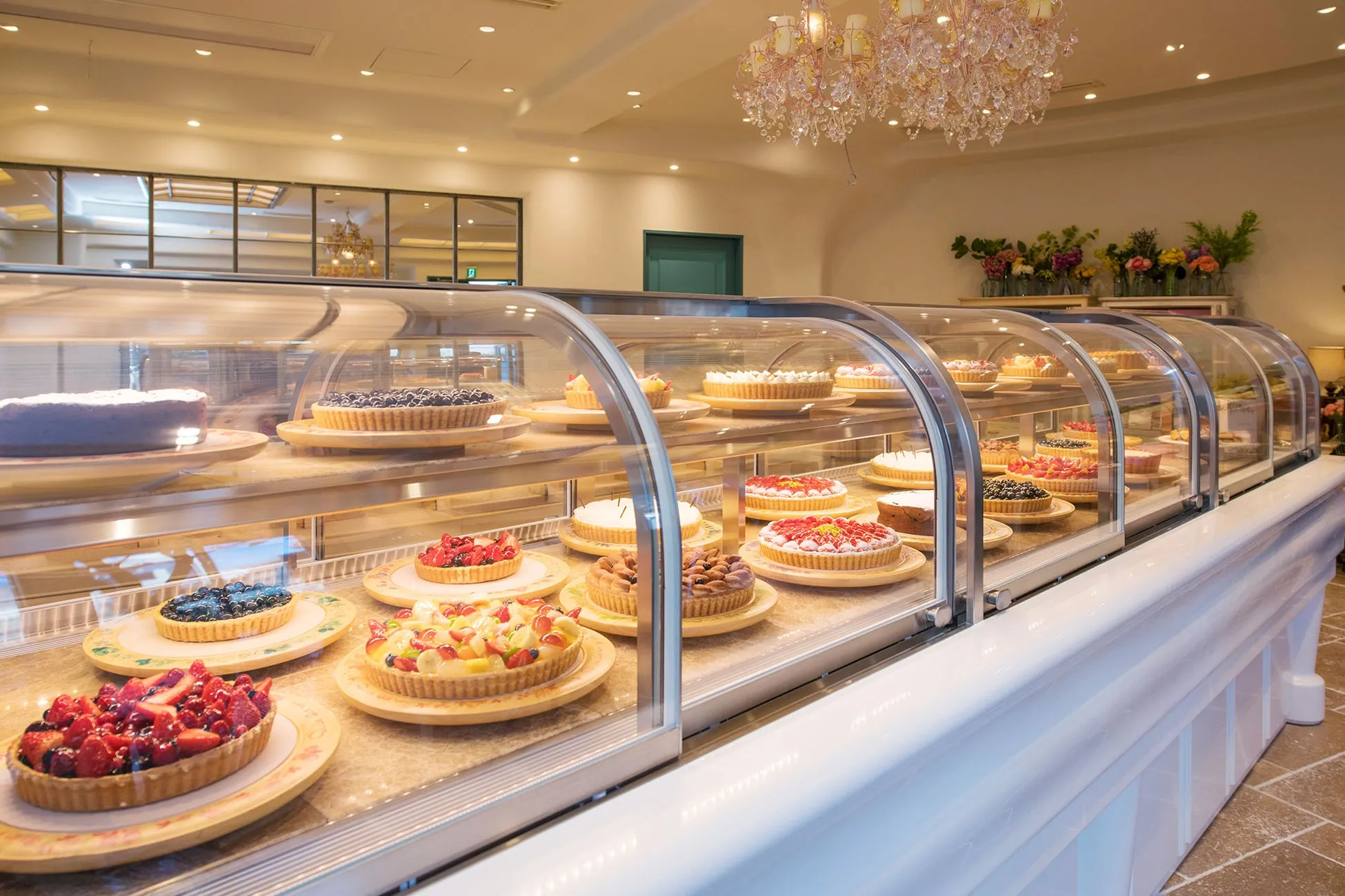 ショーケースに並んだ様々なフルーツタルトが並んだ、おしゃれなケーキ屋さんの写真です。ショーケースの上には、照明や花の装飾が施されています。