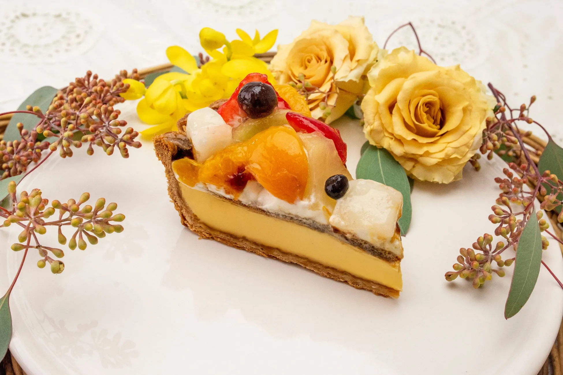 フルーツがトッピングされたタルトケーキが白い皿に盛り付けられており、黄色いバラの花と植物が添えられています。