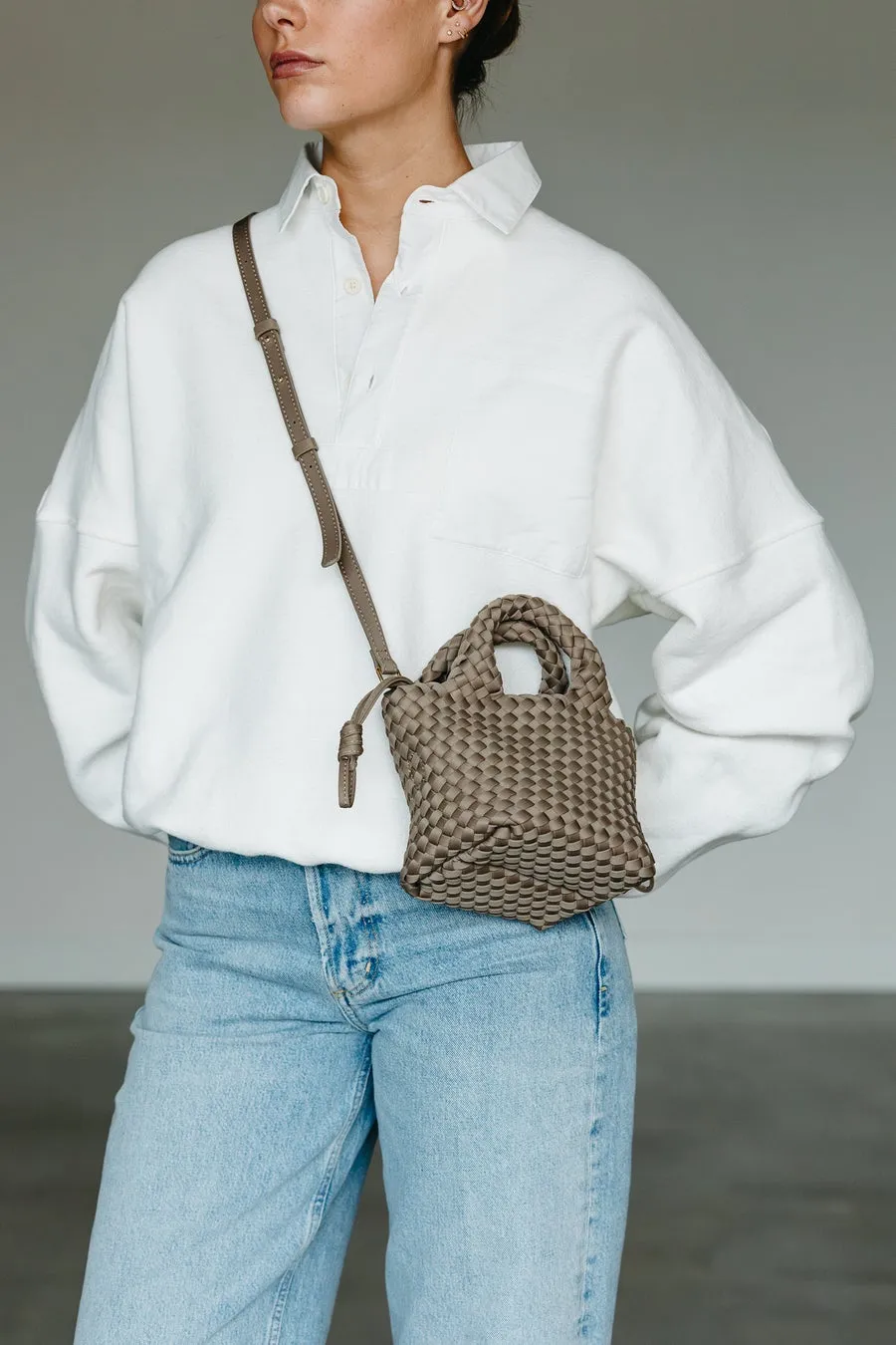 白いシャツとジーンズを着た女性が、小さな編み込みのバッグを肩にかけて立っています。