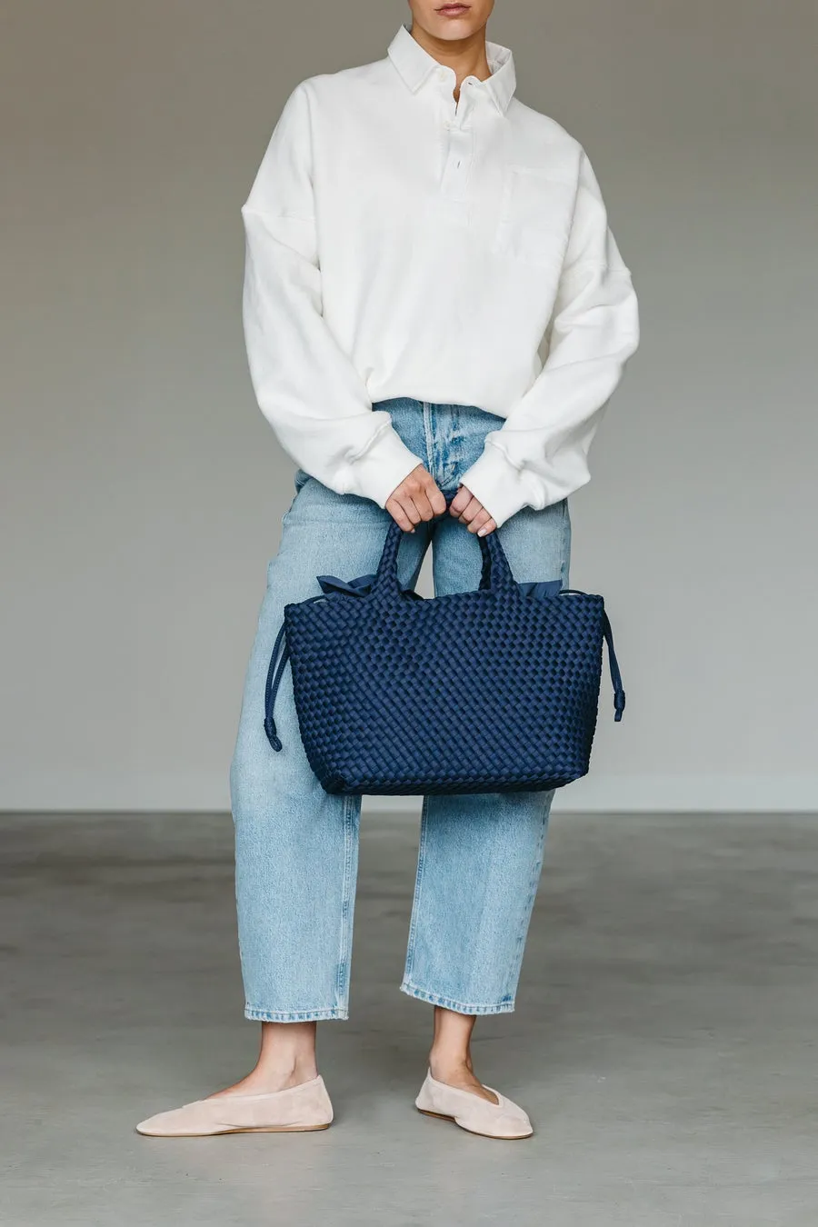 白いトップスとジーンズを着た女性が、紺色のバッグを持っています。彼女は、フラットシューズを履いています。