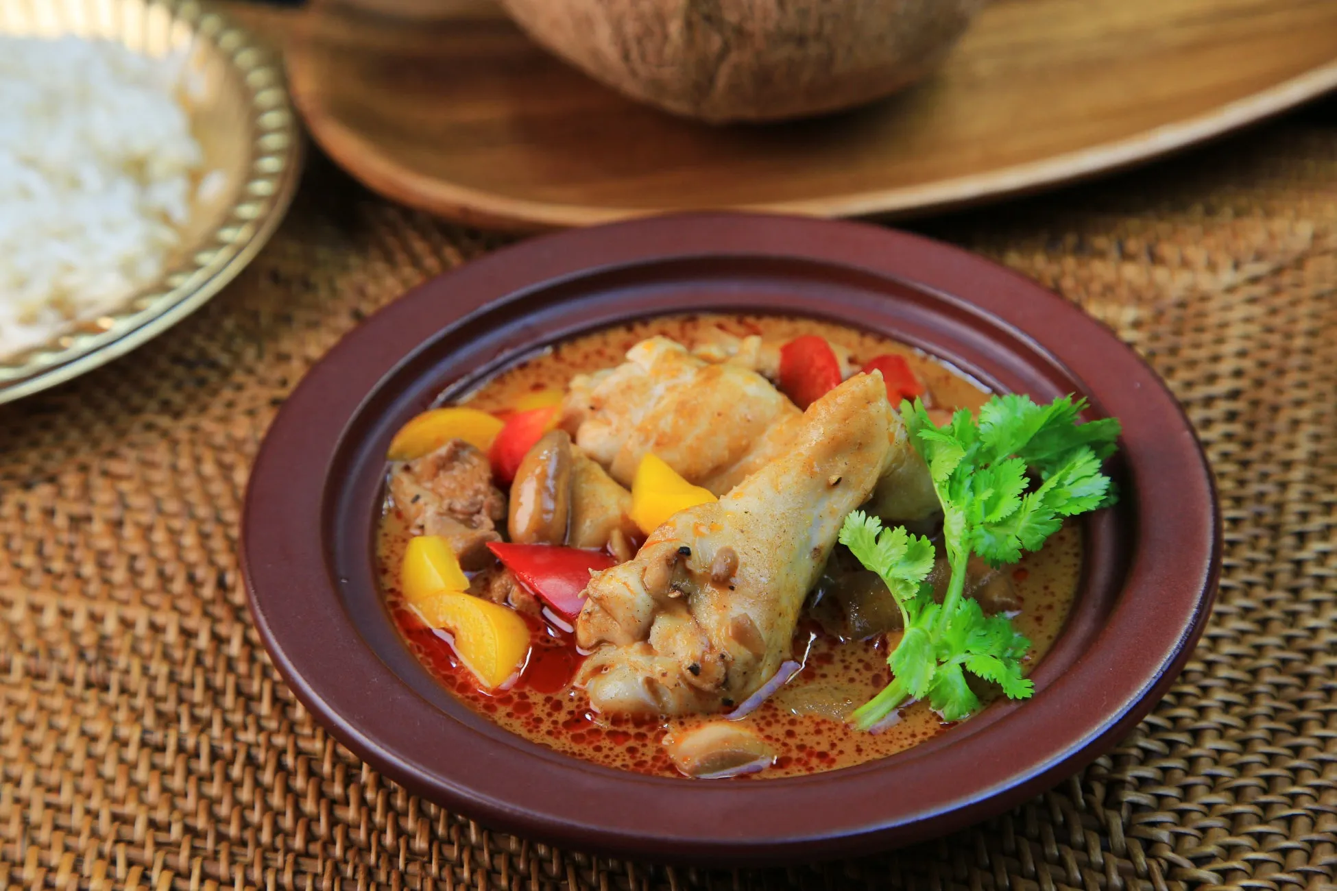 茶色の皿に入ったチキンと野菜の料理。皿のそばには米、背景にはココナッツと木製のトレイがある。