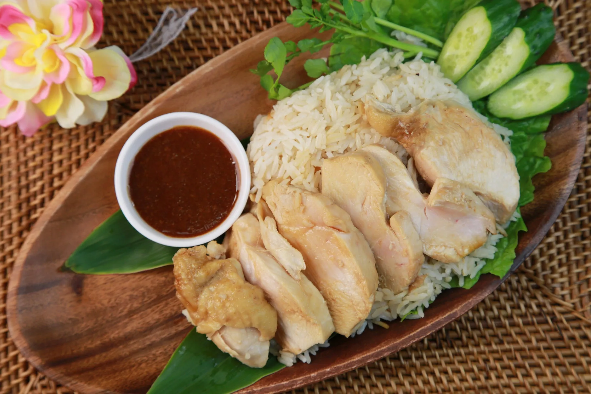 鶏肉とご飯、ソース、キュウリが木の皿に盛られた料理の写真です。背景には花と籐のようなものがあります。