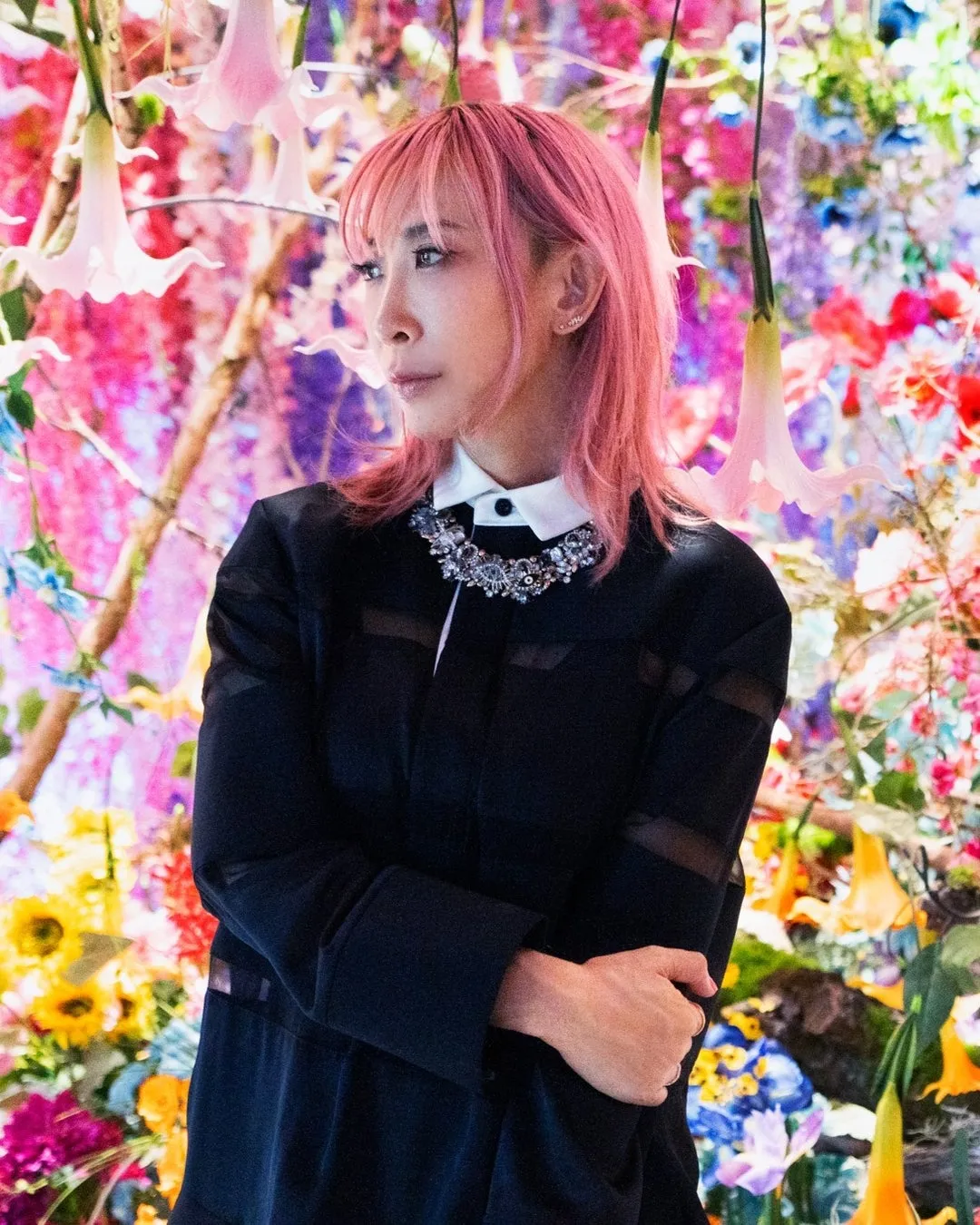 ピンク色の髪の女性が、花で飾られた背景の前に立っています。彼女は黒い服を着ており、胸元には装飾的なネックレスをしています。彼女はカメラから目をそらしており、やや遠くを見つめているようです。