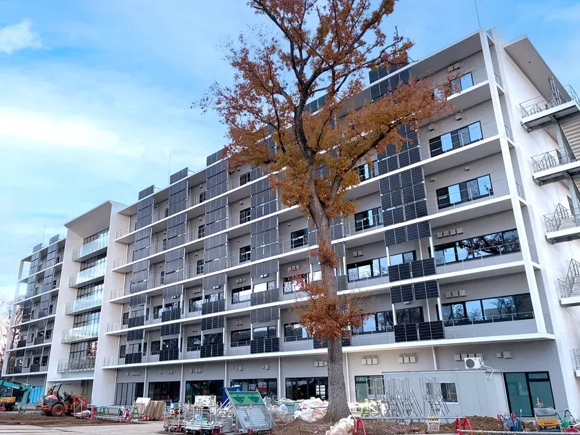 太陽光パネルが設置された集合住宅の画像です。建物の前には工事現場のような光景が広がっています。建物の横には紅葉した木が立っています。