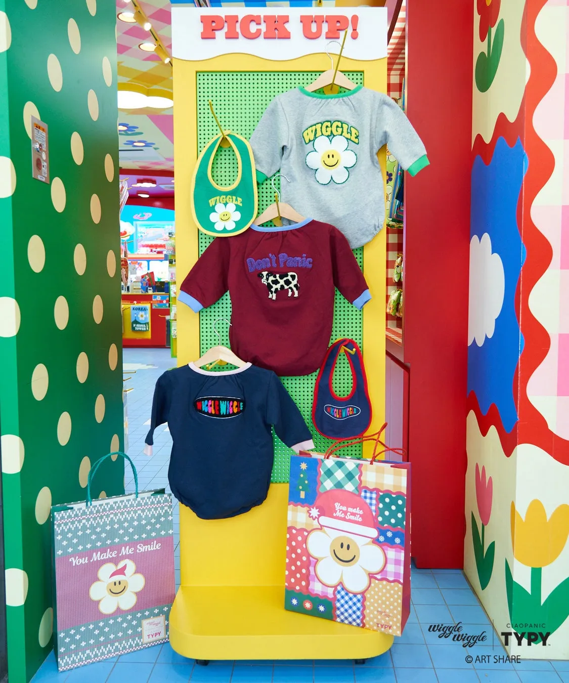 子供服とバッグがディスプレイされている写真。服には「WIGGLE」や「Don't Panic」の文字が、バッグには「You Make Me Smile」と書かれている。カラフルな背景が特徴的。