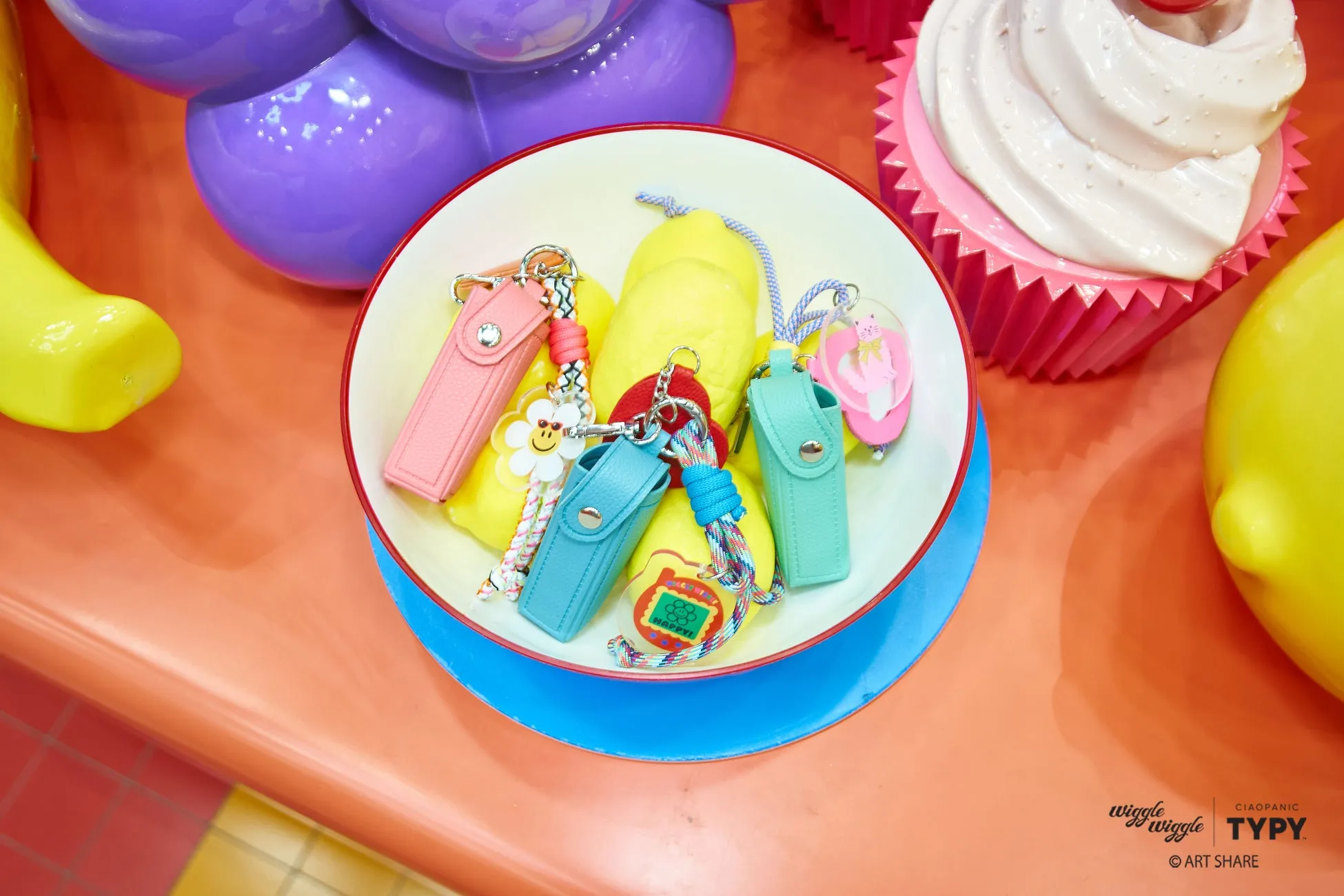 カラフルなキーホルダーとレモンが置かれた白いボウル、紫の風船、ピンクのカップケーキが写った写真。