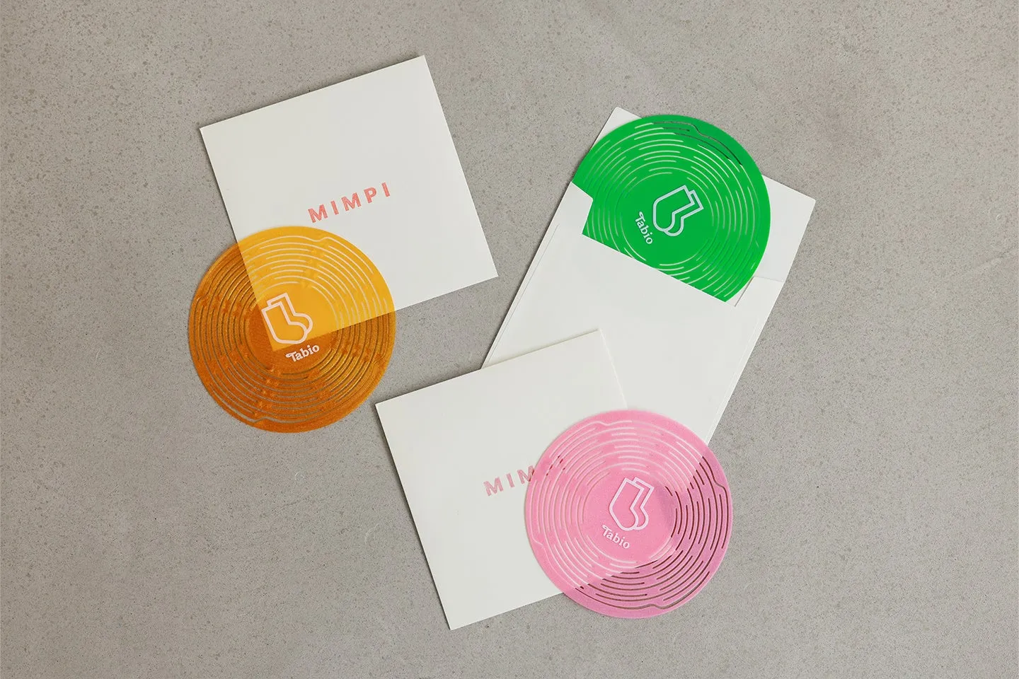 白い封筒と円形のデザインが特徴的な写真。円形デザインはオレンジ、緑、ピンクの3色で、それぞれ「Tabio」のロゴと靴下のイラストが描かれている。封筒には「MIMPI」の文字が印字されている。全体的にシンプルで洗練された印象を与える。