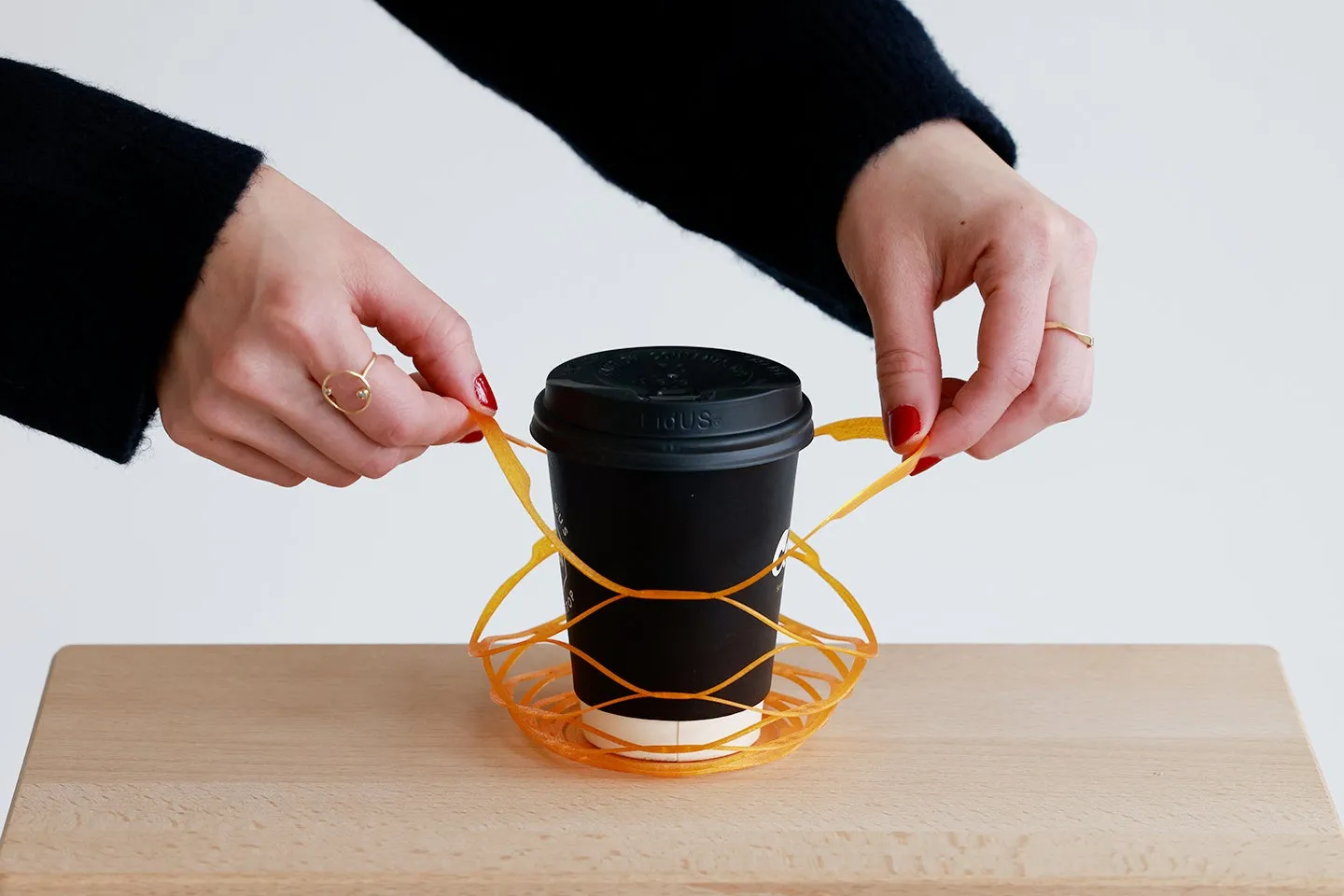 コーヒーカップをオレンジ色のホルダーに入れた様子を写した写真です。両手でホルダーの紐を引っ張っている女性が写っています。カップは黒く、ホルダーは網目状のデザインです。