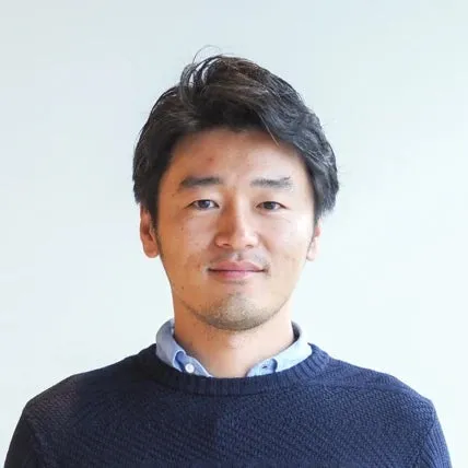 正面を向いたアジア人男性のポートレート。彼は濃い色のセーターと、その下に水色のシャツを着ている。背景は白。