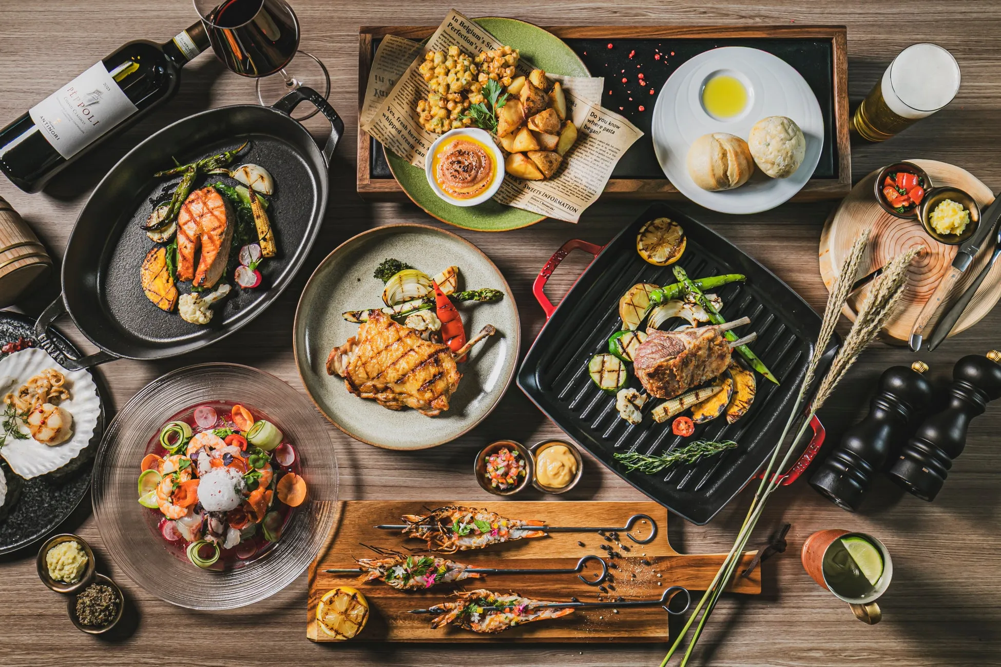 テーブルに並んだ様々な料理の俯瞰写真。グリルされた肉、魚、野菜、エビの串焼き、サラダ、パン、揚げ物、ソース、ワイン、ビールなど、様々な種類の料理が写っています。