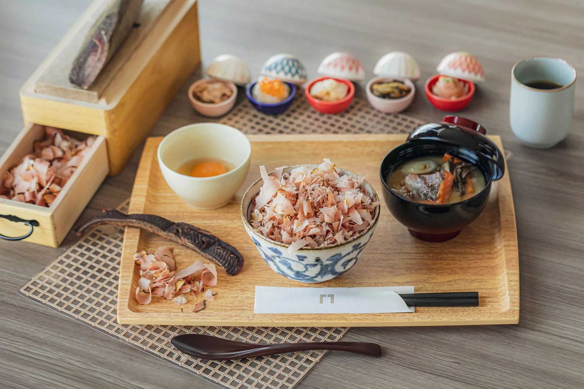鰹節と様々な小鉢が並んだ和食膳の写真。鰹節削り器、卵、味噌汁、茶碗蒸しのようなものなど、様々な料理が木製のトレイに盛り付けられています。