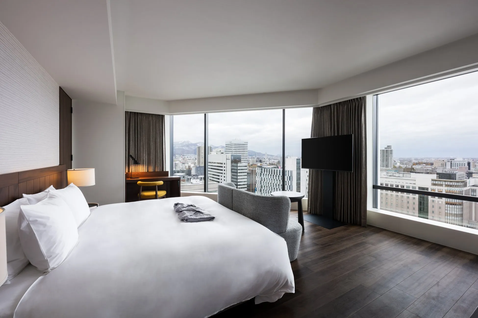 窓から街並みが見えるホテルの部屋の写真。ベッド、ソファ、机、テレビ、カーテンなどがあり、スタイリッシュな内装。