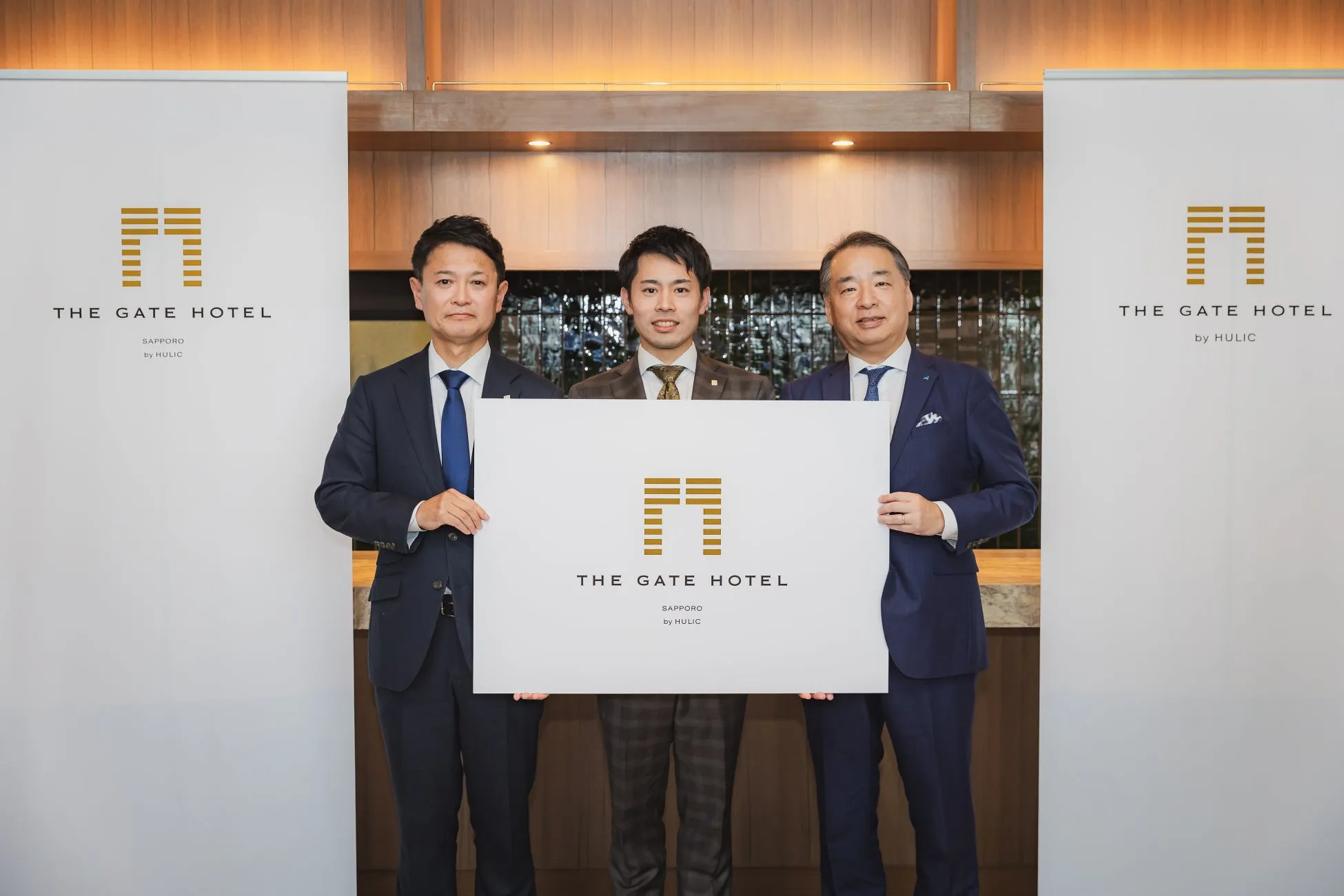 3人の男性が「THE GATE HOTEL」の看板を持って立っている写真。背景にはホテルの内装が写っている。