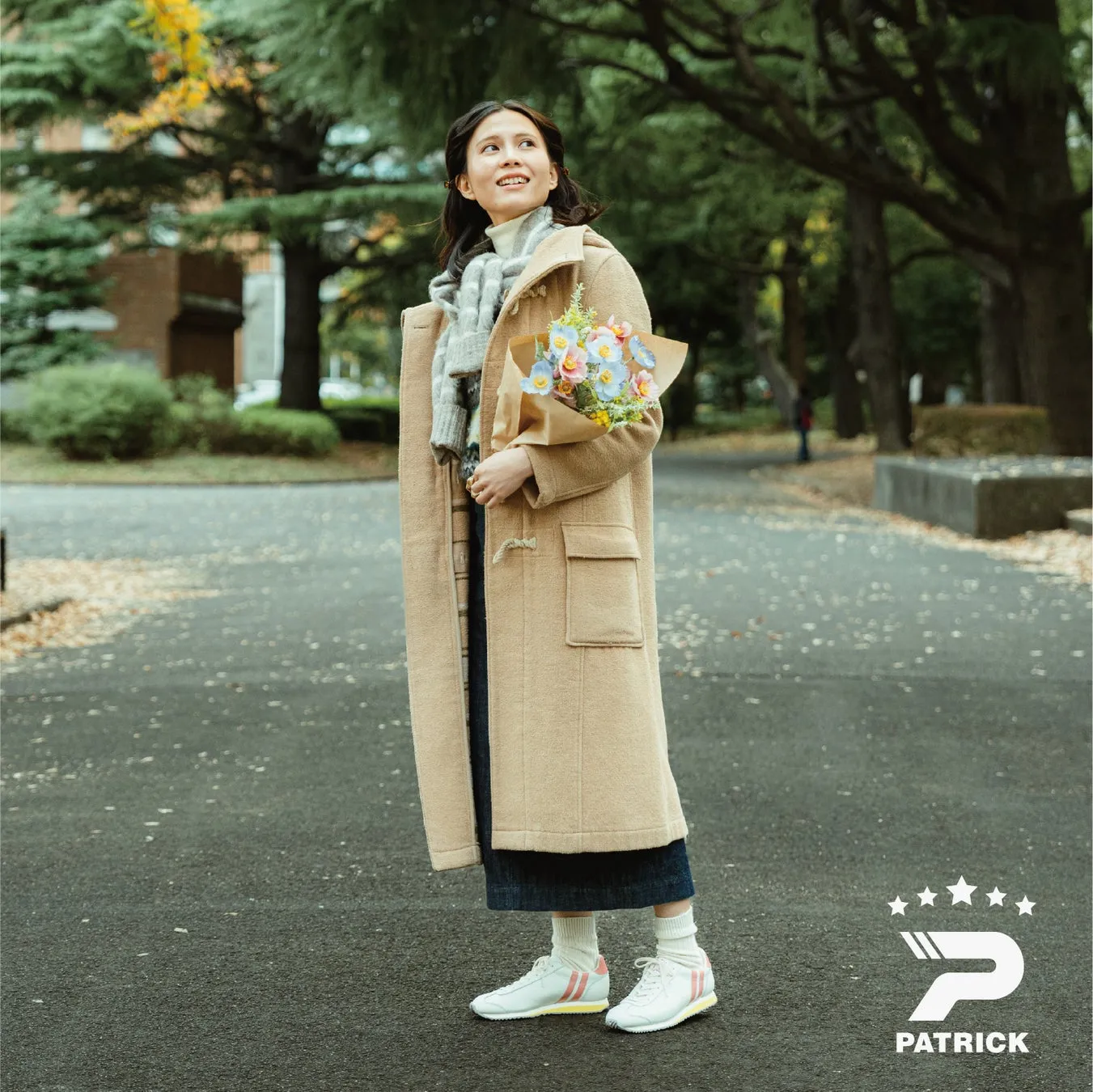 女性が花束を持って笑顔で立っている写真。ベージュのコートとスニーカーを履き、背景には木々がある。