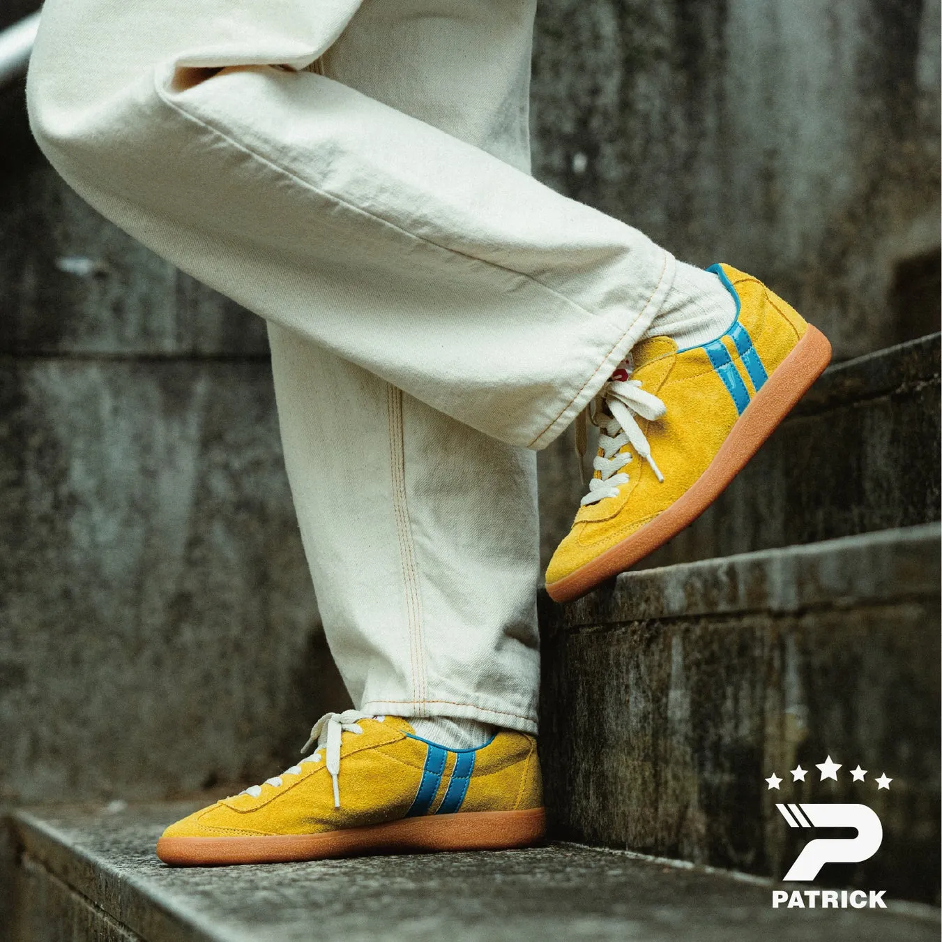 階段を上る黄色いスニーカーを履いた足元の写真。スニーカーは青いストライプが入っており、靴底は茶色、白いズボンを履いている。