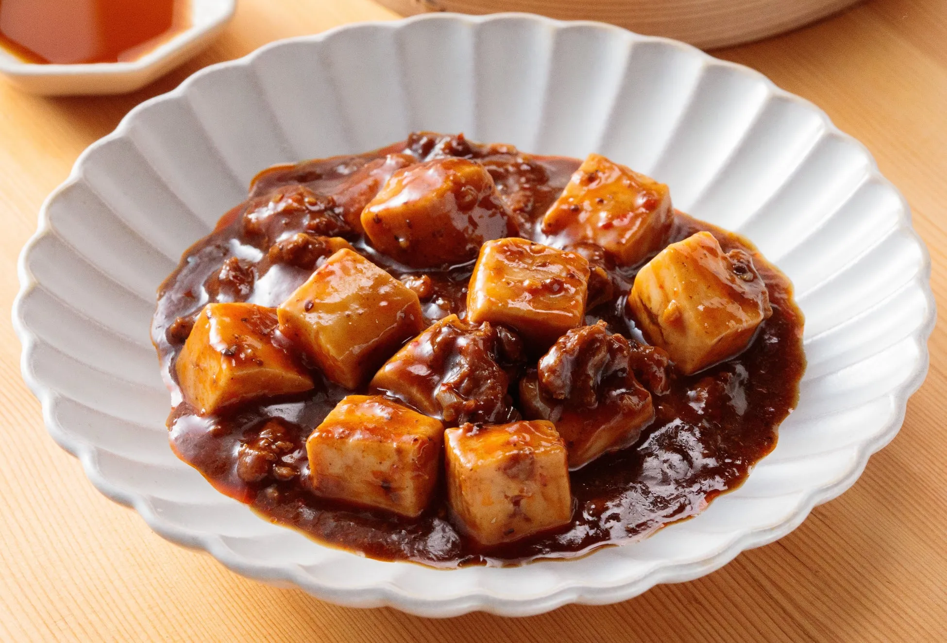 白い皿に盛られた麻婆豆腐のクローズアップ写真。豆腐は立方体にカットされ、茶色のソースで覆われています。背景には、木製の表面と小さなソース皿があります。