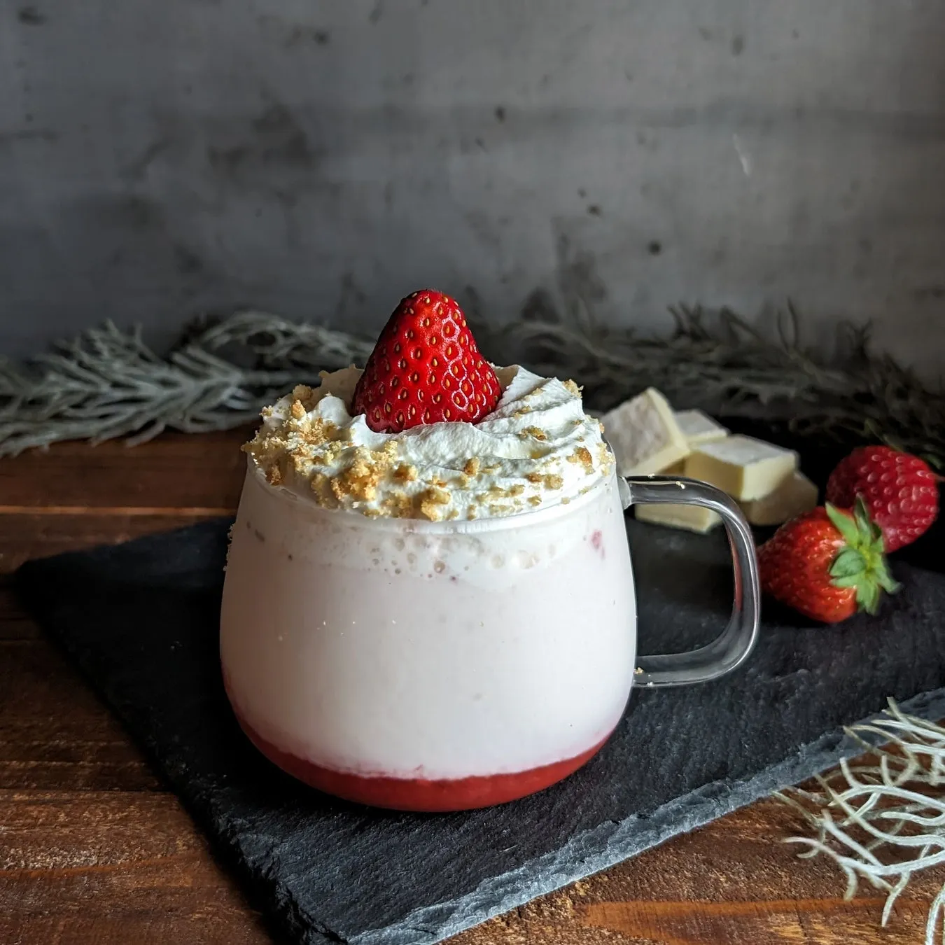 いちごがトッピングされた、クリームと砕いたクッキーで飾られたピンク色のドリンクのクローズアップ写真。ドリンクは透明なガラスのマグカップに入っていて、黒い石板の上に置かれています。背景には、白いチョコレートと、さらにいちごが添えられています。