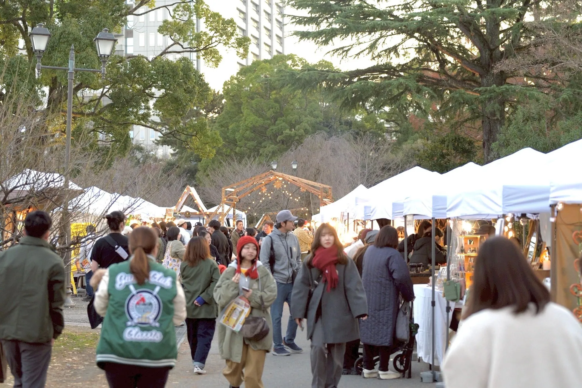 公園で開催されているマーケットの様子を捉えた写真です。多くの人々が屋台を訪れ、楽しんでいます。木々や建物も写っており、賑やかな雰囲気が伝わってきます。