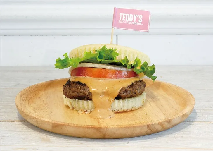 木製の皿に載ったハンバーガーの写真。 ハンバーガーは、レタス、トマト、玉ねぎ、ソース、パティ、バンズで構成されています。 ハンバーガーには、TEDDY'S BIGGER BURGERSと書かれた小さなピンク色の旗が刺さっています。