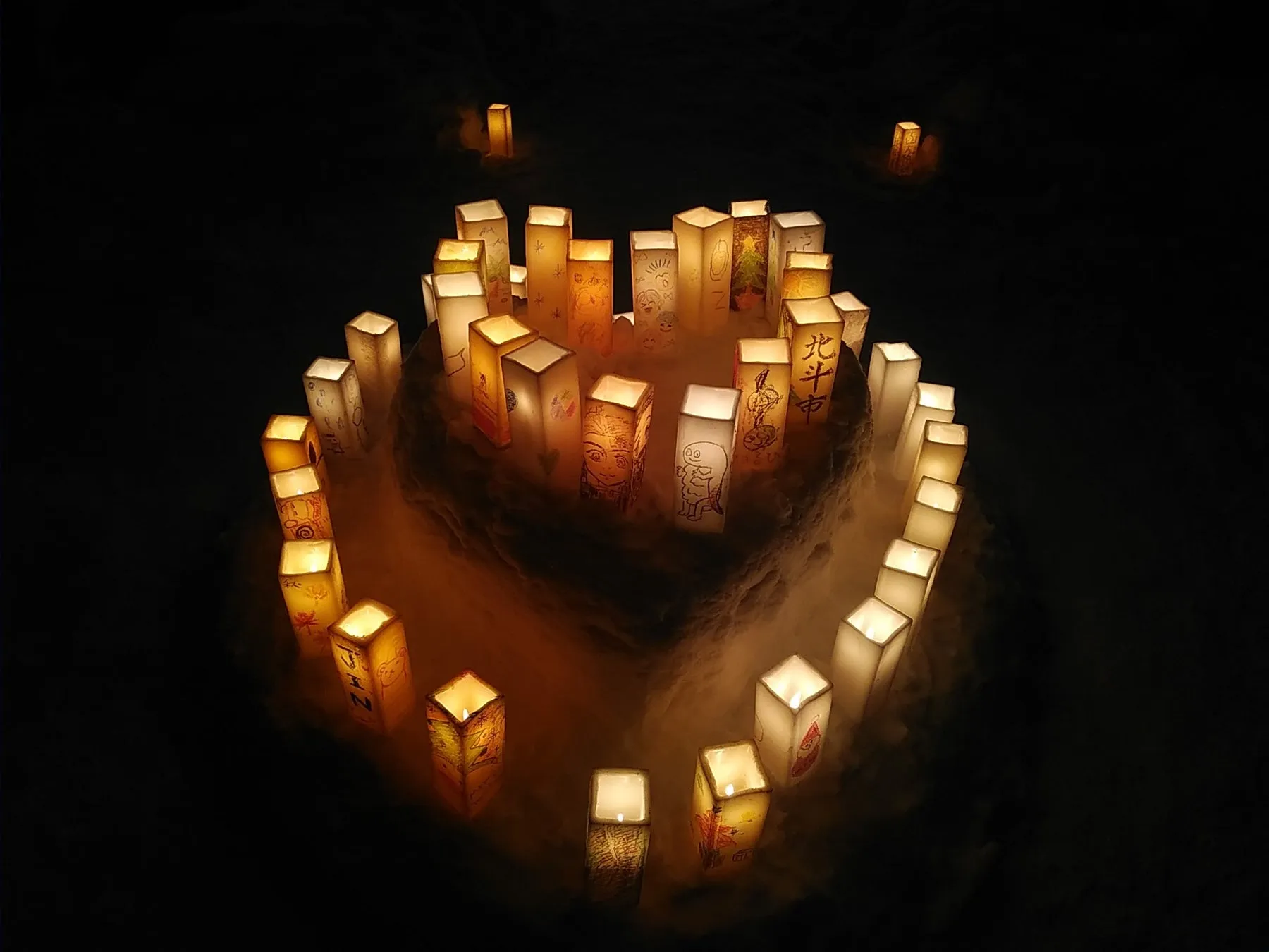 雪の中に置かれた多数の灯篭の写真。灯篭は四角い形で、それぞれに絵や文字が描かれている。灯篭はハートの形に配置され、幻想的な雰囲気を作り出している。