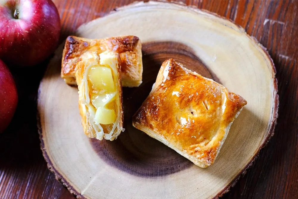 木製のまな板の上に、アップルパイとリンゴが置かれた写真です。アップルパイは焼き色が良く、中にはリンゴが詰まっています。リンゴは赤く、美味しそうです。
