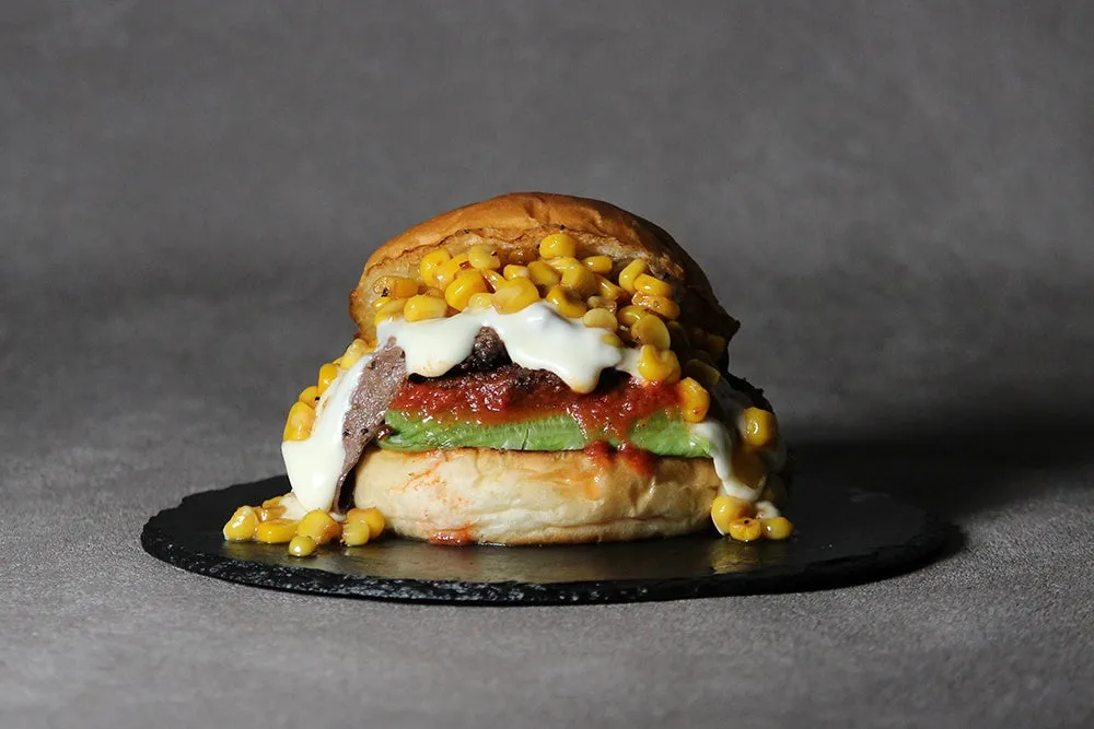 トッピングがたっぷりのハンバーガーの写真。バンズの間にはアボカド、ミートパテ、ソースが挟まれ、上にはコーンが山盛りになっている。