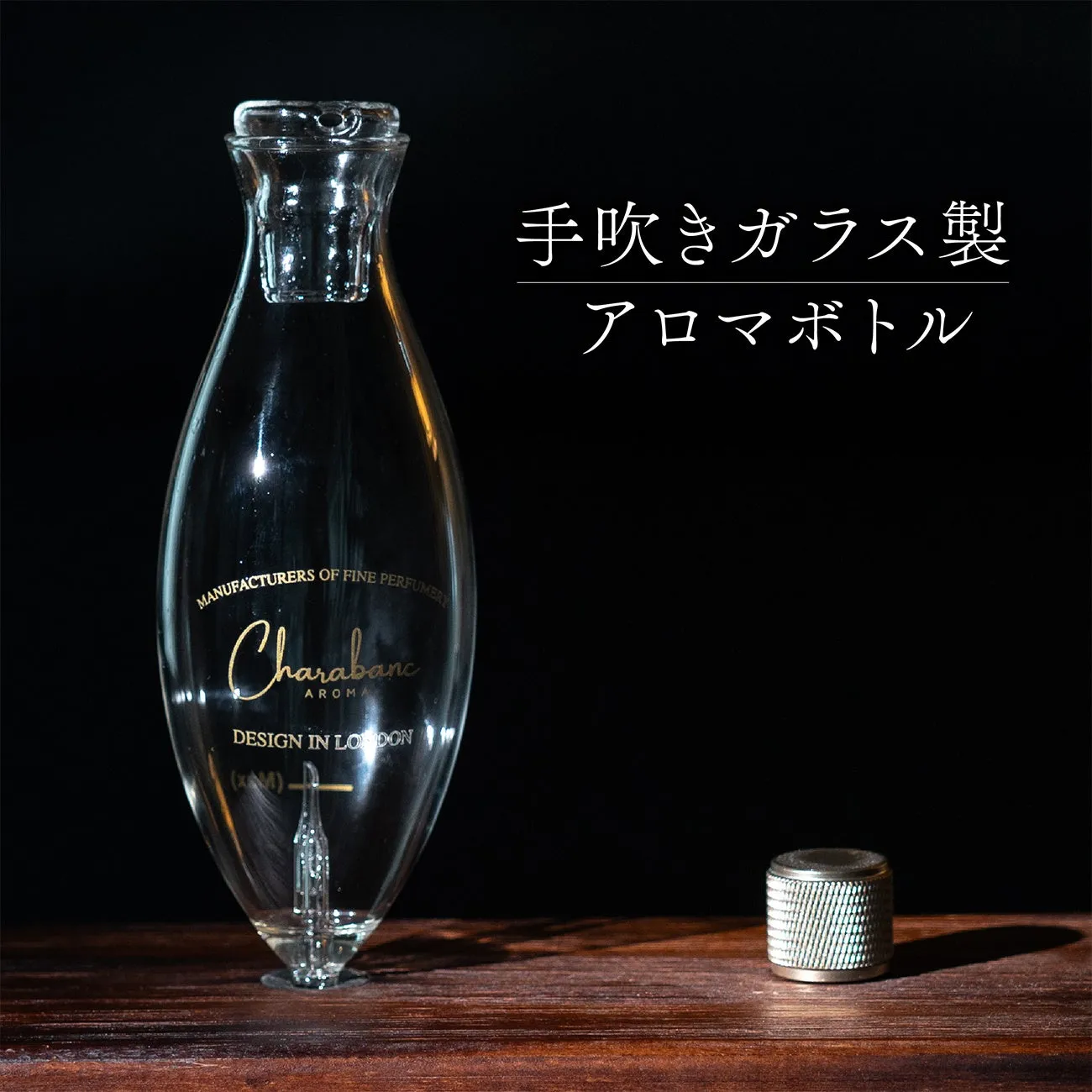 透明なガラス製のアロマボトル。手吹きガラス製で、ロゴとデザインが施されている。横には蓋が置かれている。