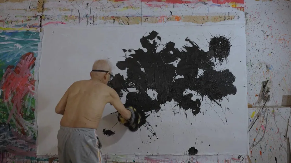 画家が大きなキャンバスに黒い絵の具で抽象的な絵を描いている様子。