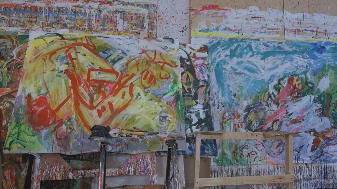 絵画が展示されたアトリエの風景。様々な色と形の抽象的な絵画が壁に立てかけられ、画材や小物が置かれたテーブルが手前に見える。