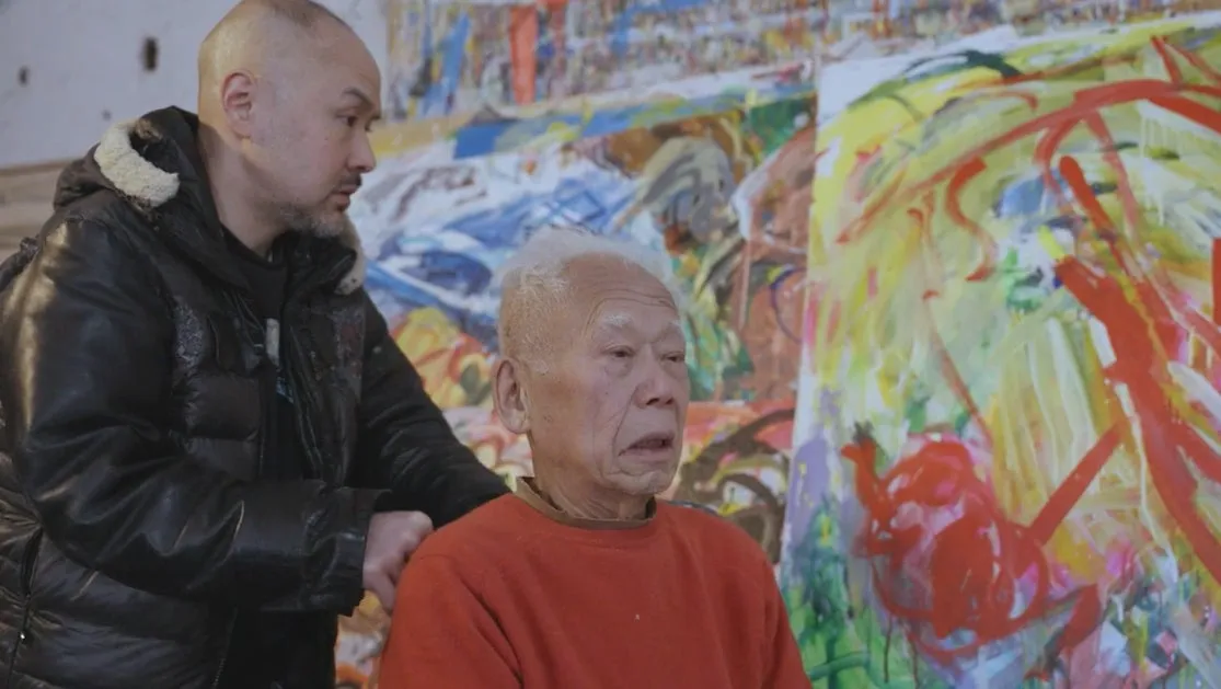 2人の男性が、絵画の前で立っている写真。一人は若い男性で、黒いジャケットを着ています。もう一人は年配の男性で、オレンジ色のセーターを着ています。背景には抽象的な絵画があります。