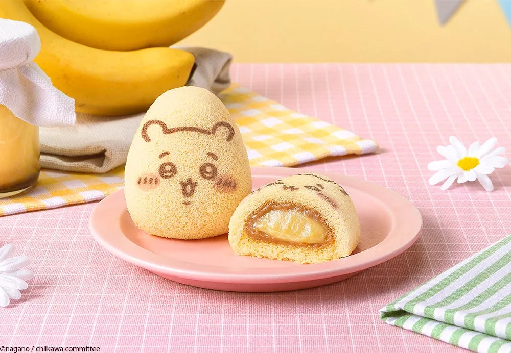 かわいらしいキャラクターのケーキがピンク色の皿に載せられ、バナナや花、チェック柄の布と一緒に配置されている写真です。