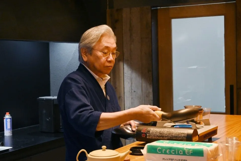 男性が調理器具を使って何かを作っている様子が写っています。彼はメガネをかけており、真剣な表情をしています。テーブルの上には、ティーポット、ティッシュボックス、ガスコンロなどがあります。