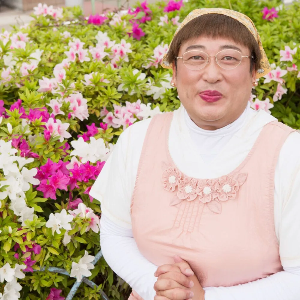 花の前でポーズをとる男性。彼はピンクの衣装を着ており、メガネをかけています。