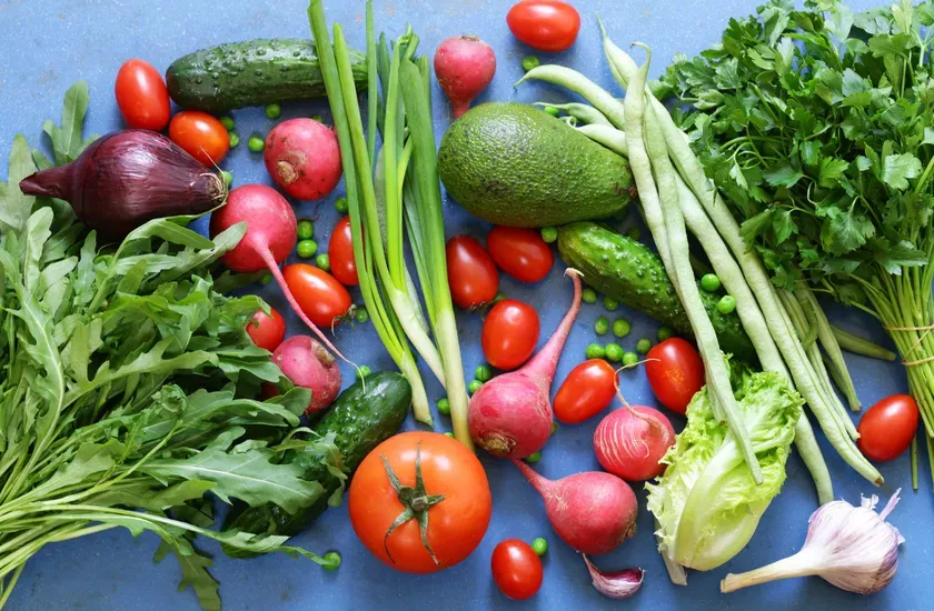 様々な野菜が青い背景に配置された写真です。トマト、ラディッシュ、アボカド、キュウリ、ネギ、インゲン、パセリ、レタス、エンドウ豆、ニンニク、紫玉ねぎなどの野菜が含まれています。
