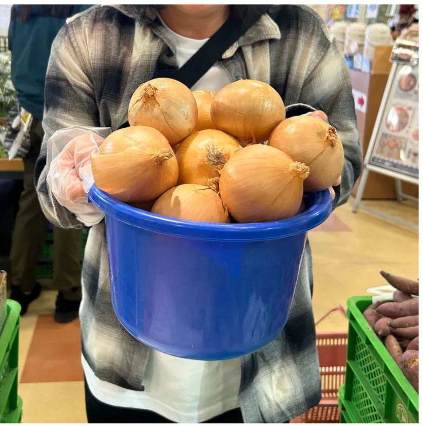 人が玉ねぎの入った青いバケツを持っている写真です。バケツにはたくさんの玉ねぎが入っており、人がそれらを持ち上げています。背景には、他の野菜が入った緑色のバスケットや、他の人々がいるのが見えます。