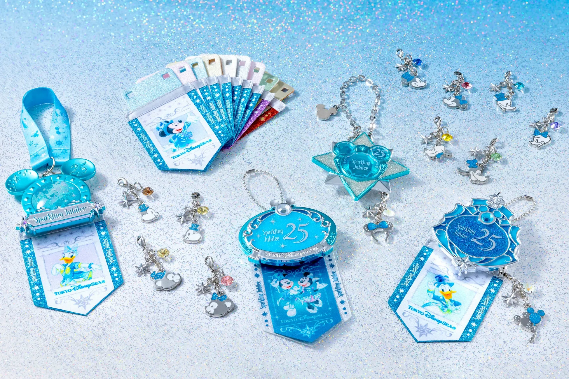 東京ディズニーシー25周年記念のキラキラしたアクセサリーの集合写真。ミッキー、ミニー、ドナルド、デイジーなどのキャラクターが描かれたタグやチャーム、キーホルダー、ストラップなどが写っています。背景は水色で、キラキラとした光が散りばめられています。
