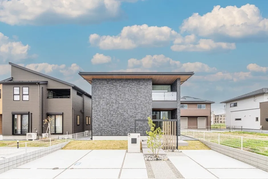 モダンなデザインの住宅の外観写真です。空は青く、白い雲が浮かんでいます。住宅はグレーの石壁と木材が組み合わされており、スタイリッシュな印象を与えます。庭には小さな木が植えられており、洗練された雰囲気を醸し出しています。
