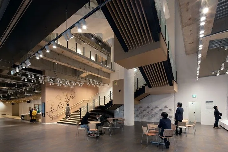 モダンな内装の建物で、人々が椅子に座って会話したり、階段を上がったりしている様子